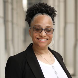 Terry Harris Headshot where she is wearing a white blouse, long silver necklace, black blazer, and black framed glasses. She is wearing her hair up in a high short pony tail.