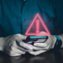 Person typing on phone with red warning symbol floating above the phone.